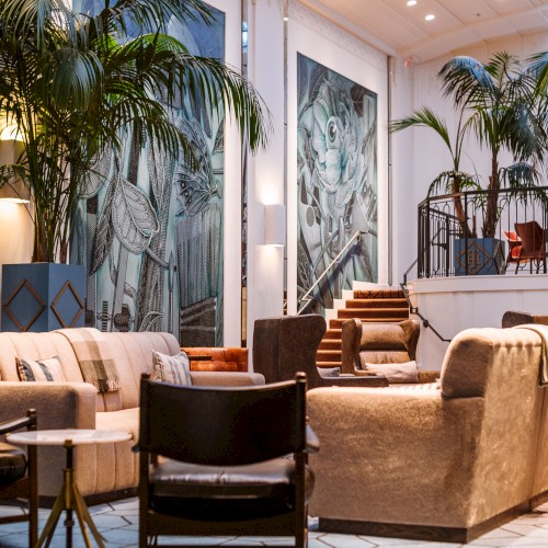 A chic, lush lounge with tall palm plants, modern beige sofas, and dark wood chairs; bright art panels and soft lighting create a tropical, upscale vibe.