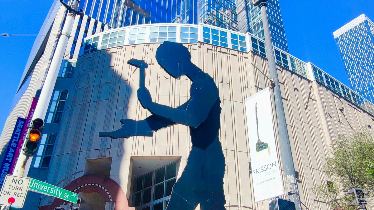 A large blue silhouette sculpture of a basketball player dunking outside a modern glass building, with pedestrians on a sunny street.