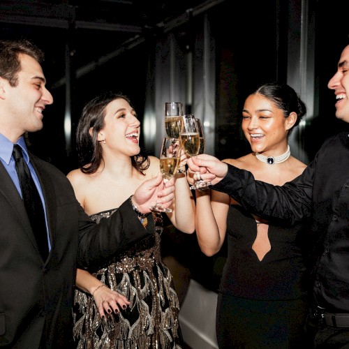 A group of four adults in formal attire clinking champagne glasses at a celebration. Their smiles and toasts show festive mood, dim background.