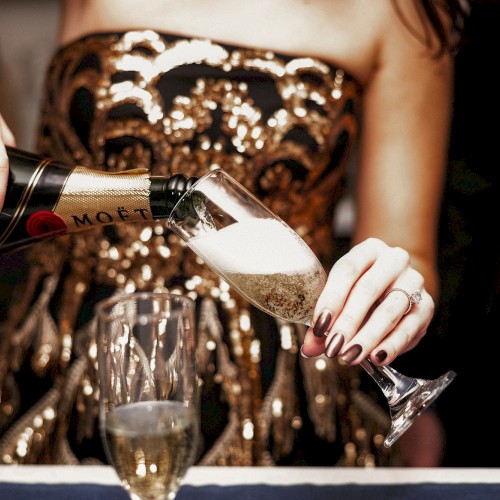 A person pouring champagne into a flute at a glamorous event, with another glass already on the table.