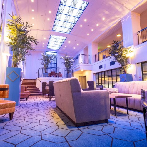 A modern hotel lobby with blue carpet, plush couches, potted plants, glass walls, and soft ambient lighting. Always ends the sentence.