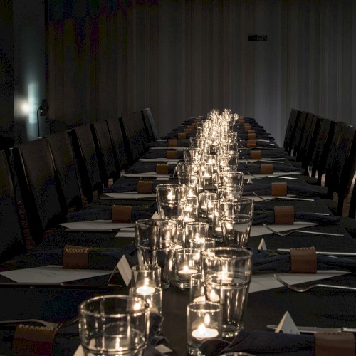 A long, dimly lit dining table with candles, place settings, and napkins arranged neatly along both sides in a formal event setup.