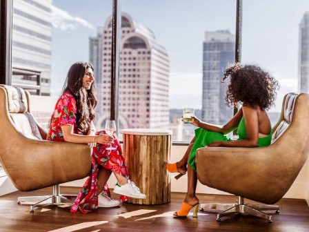 Two women chat in a bright, modern lounge by large windows, sitting in tan chairs with a wooden table between them, city skyline behind.