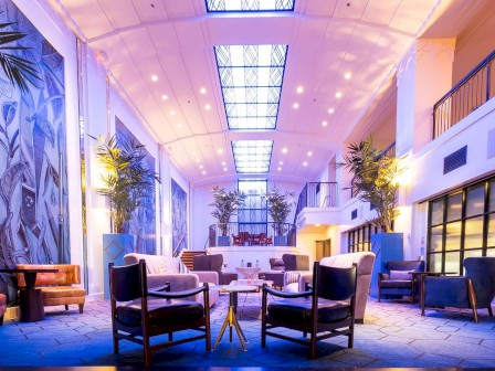 A bright, modern hotel lobby with high ceilings, tropical wall mural, potted plants, and stylish seating around small tables.