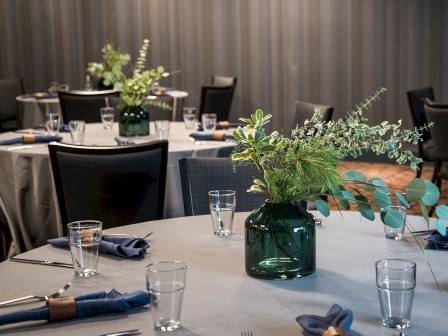 The image shows a dining setup with round tables draped in white tablecloths, green glass vases with greenery, and blue napkins, in an elegant setting.