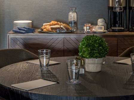A round table with glasses, notepads, and a potted plant; behind it, a buffet with plates, pastries, and a coffee station ending the sentence.