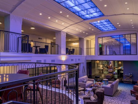 A modern hotel lobby with a mezzanine, lounge seating, glass walls, and blue-purple lighting.