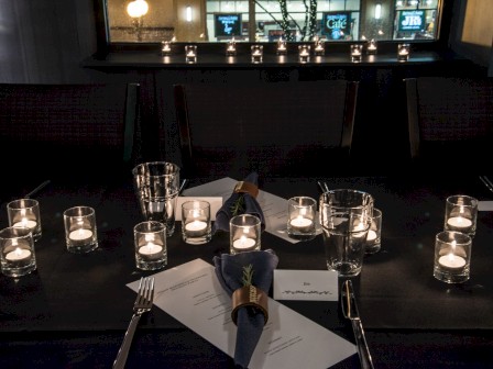 A dimly lit dining setup with numerous candles, glasses, napkins, cutlery, and menus arranged on a dark table in a cozy restaurant setting.