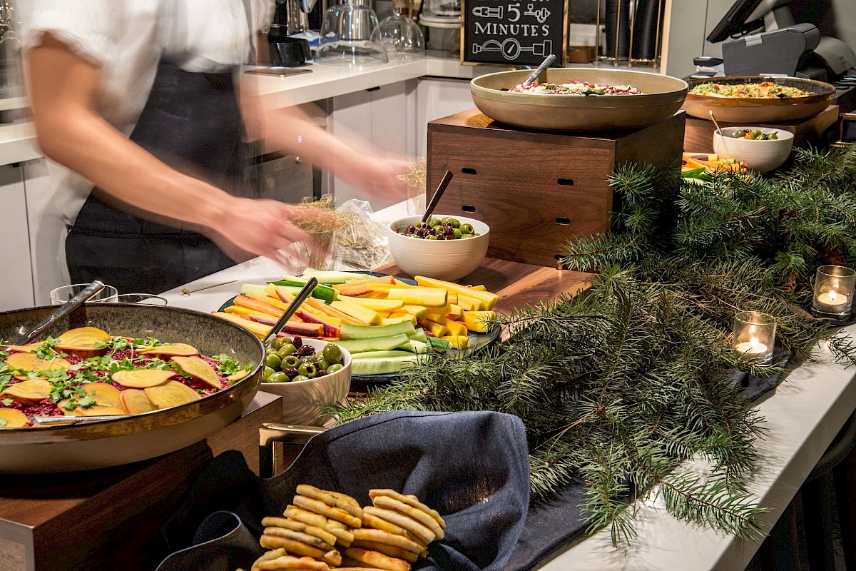 A chef arranges fresh dishes on a counter with bowls, veggies, and herbs; plates of bruschetta, olives, and sliced fruit on display.