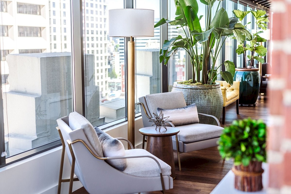 A modern living space with light gray chairs, a small round table, a tall lamp, large windows, and lush green plants.