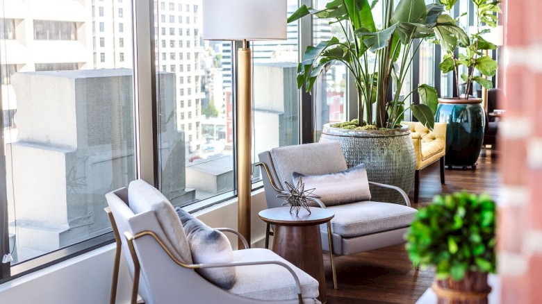 A modern living space with light gray chairs, a small round table, a tall lamp, large windows, and lush green plants.