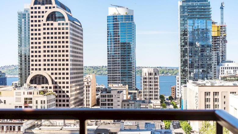 A city skyline with modern glass towers rises over lower buildings, seen from a balcony railing against a clear blue sky.