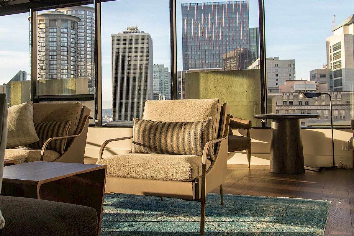 A modern living room with beige sofas, throw pillows, and glass walls overlooking a city skyline with tall buildings, a cozy rug, and a coffee table.