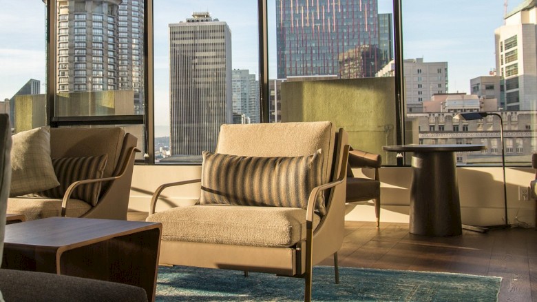 A modern living room with beige sofas, throw pillows, and glass walls overlooking a city skyline with tall buildings, a cozy rug, and a coffee table.