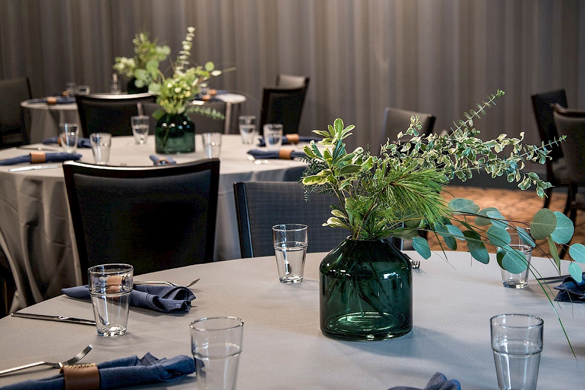A banquet hall with round tables, blue napkins, clear glasses, and green leafy centerpieces in dark glass vases.