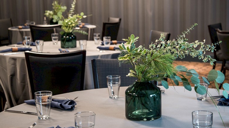 A banquet hall with round tables, blue napkins, clear glasses, and green leafy centerpieces in dark glass vases.