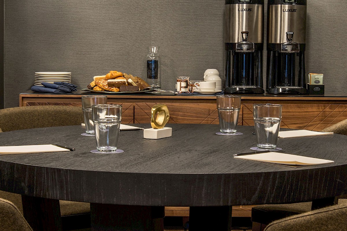 A round conference table set with glasses and napkins, a coffee station with carafes and snacks in the background.