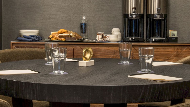 A round conference table set with glasses and napkins, a coffee station with carafes and snacks in the background.