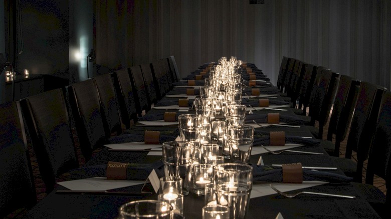 A long dining table set for a formal event, with candles, glassware, and place settings along both sides in a dim, intimate room.