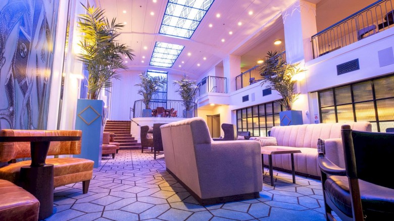 A modern hotel lobby with blue carpeting, plush seating, potted plants, and high ceilings; warm lighting and a welcoming, airy vibe.