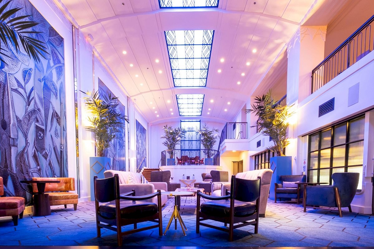 A bright, modern hotel lobby with tall plants, blue-toned lighting, and stylish seating around small tables; a calm, inviting space.