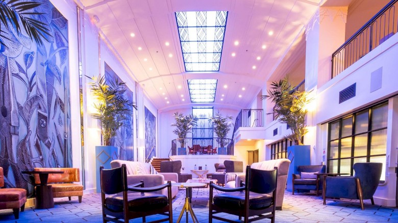 A bright, modern hotel lobby with tall plants, blue-toned lighting, and stylish seating around small tables; a calm, inviting space.