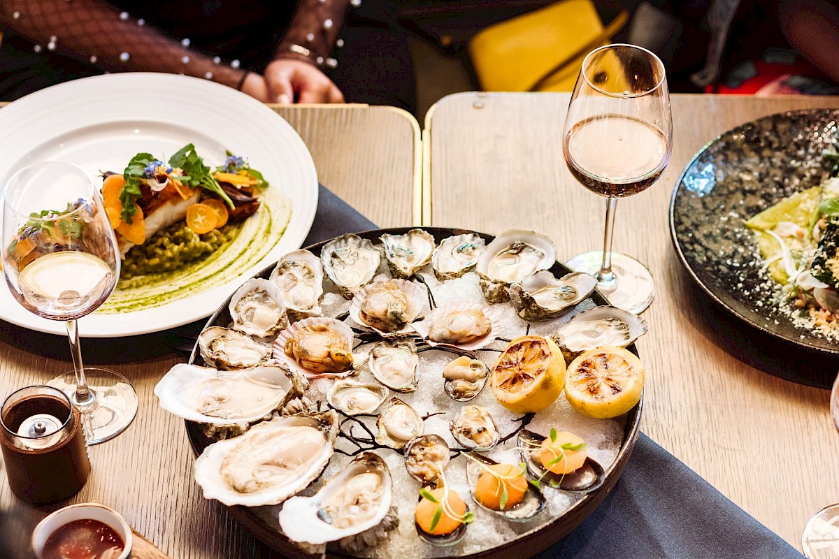 A table of oysters on a platter with lemon wedges, wine glasses, a plate of colorful food, and drinks in a cozy dining setting.