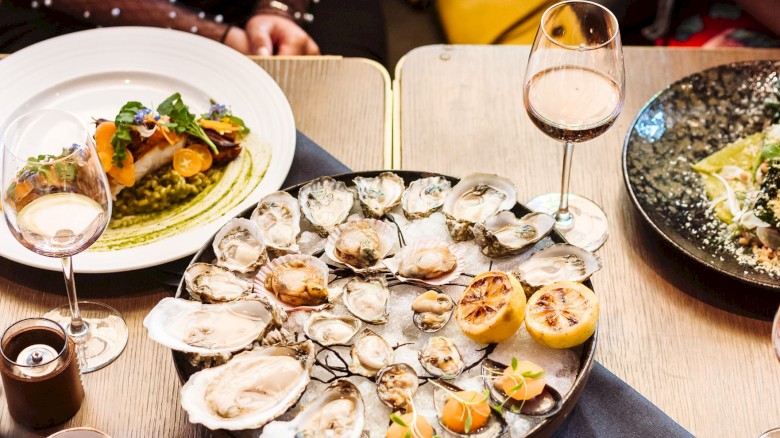 A table of oysters on a platter with lemon wedges, wine glasses, a plate of colorful food, and drinks in a cozy dining setting.