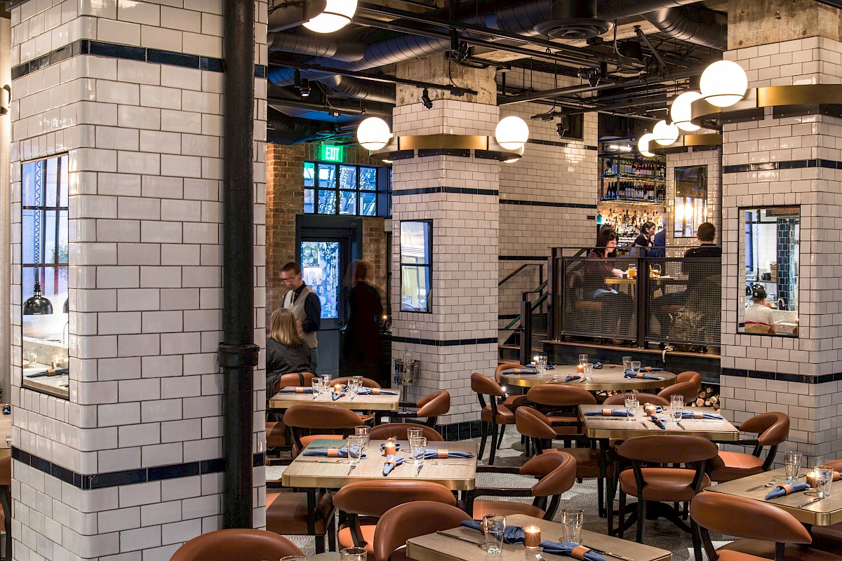 A stylish, retro-style restaurant with white tile walls, warm lighting, and brown chairs; tables set with cutlery and glassware, an inviting dining hall.