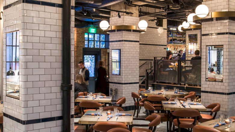 A stylish, retro-style restaurant with white tile walls, warm lighting, and brown chairs; tables set with cutlery and glassware, an inviting dining hall.