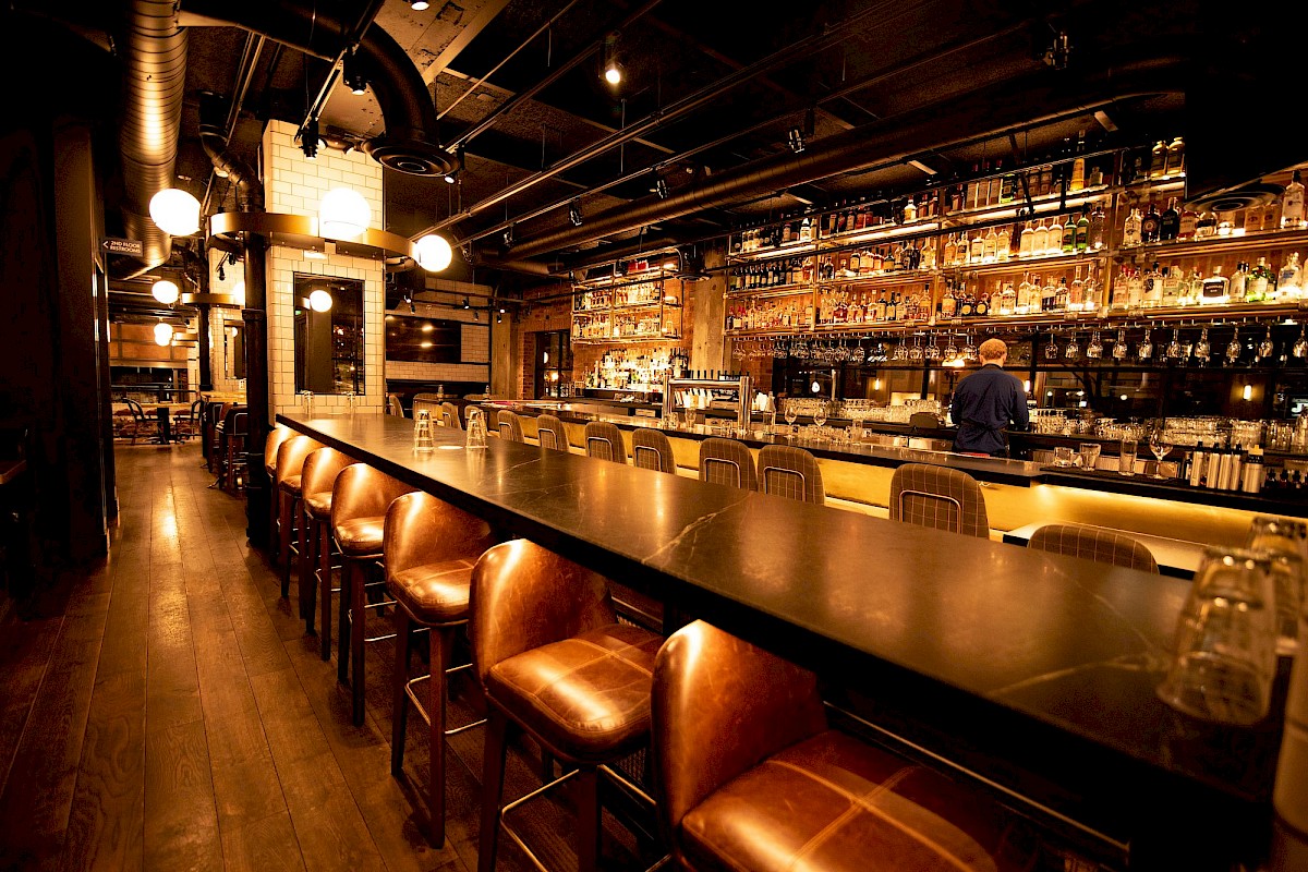 A long, dark bar with leather stools, bottles on shelves, warm lighting, and a person bartending in the background.