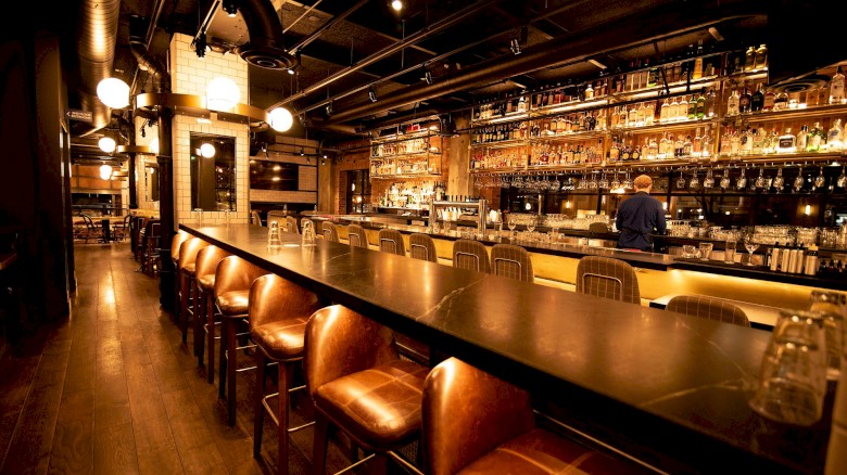 A long, dark bar with leather stools, bottles on shelves, warm lighting, and a person bartending in the background.