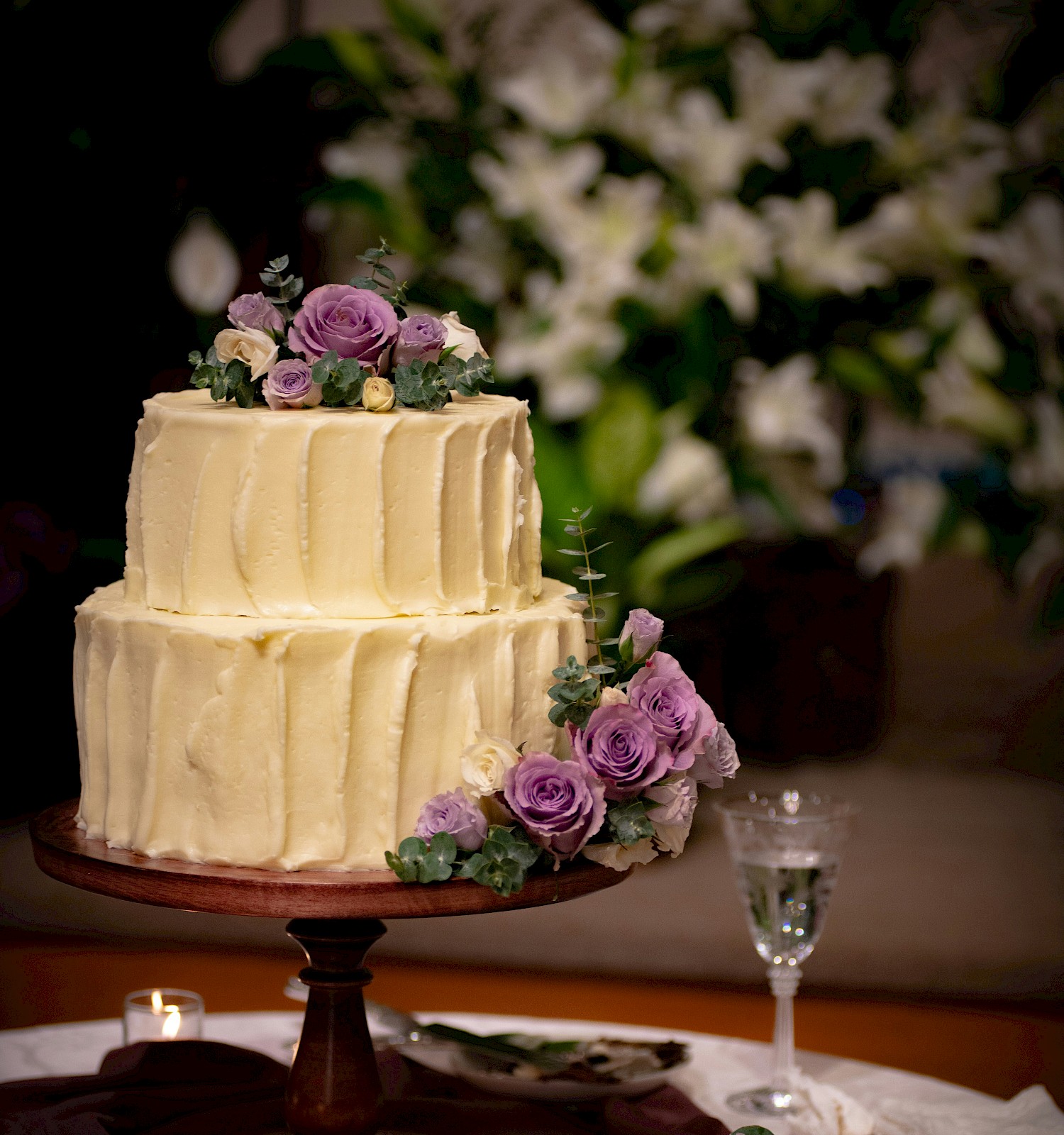 A two-tier cream cake with purple flowers on top and side, on a wooden stand, with candles and a floral backdrop.