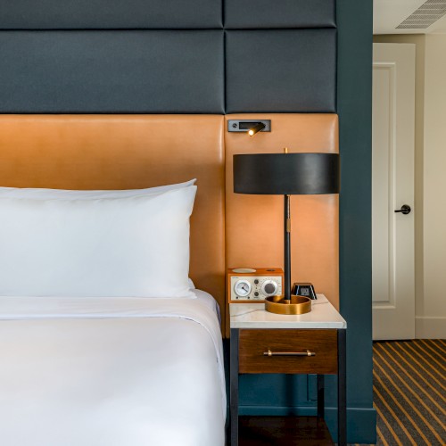 A stylish hotel room with a white bed, orange headboard panel, a nightstand with a lamp, and a radio, beside a doorway in a tidy space.