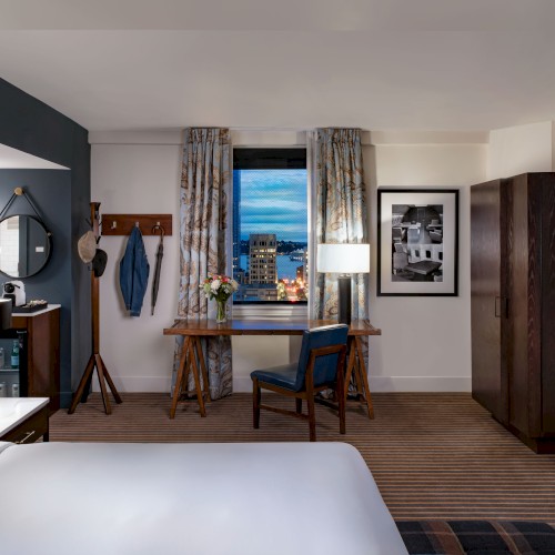 A hotel room with a bed, desk by a window, a wardrobe, a chair, lamps, wall art, and patterned curtains—cozy and modern with city view.