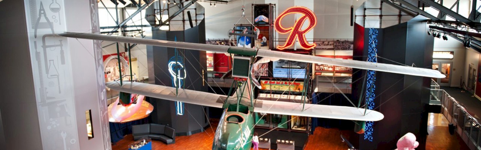 An indoor toy showroom with model airplanes suspended from the ceiling, colorful displays, a pink ride-on car, and a large red “R” logo in the background, viewed from above.