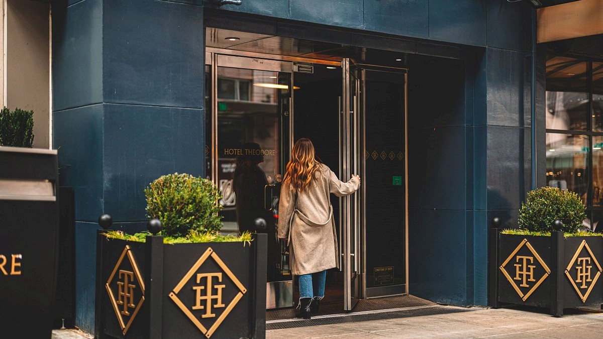 A woman enters a stylish blue building through a glass door, stepping onto a street lined with decorative planters and geometric logos, at address 1531.