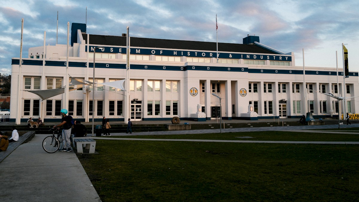 A white, historic-looking museum building with a blue stripe, large windows, and people strolling outside near a green lawn and benches.