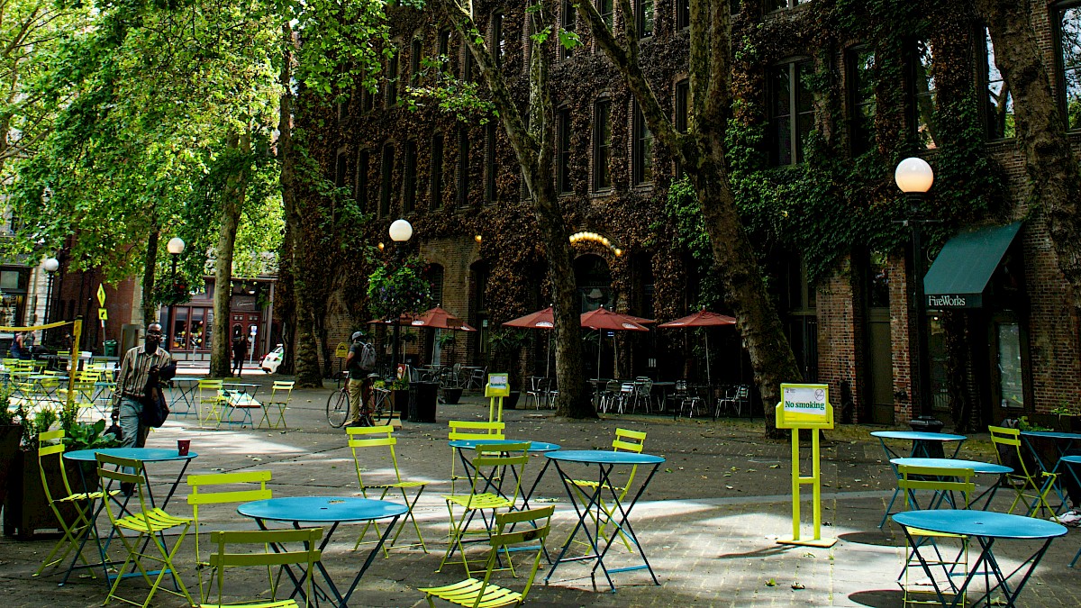A tree-lined outdoor cafe with colorful metal tables and chairs, shaded by tall green-leafed trees along a brick building street.
