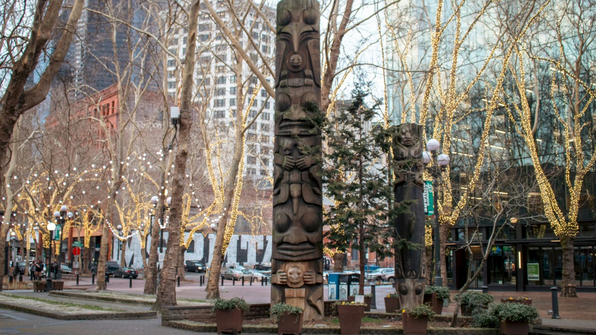 Two tall carved totems in a park with leafless trees, urban background, people strolling, holiday lights, and potted plants nearby.