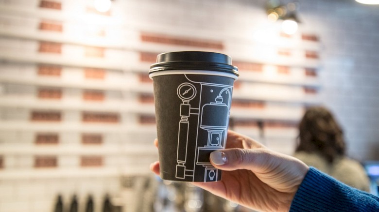 A hand holds a coffee cup with a design, in a cafe setting featuring a blurred brick and tile wall in the background.