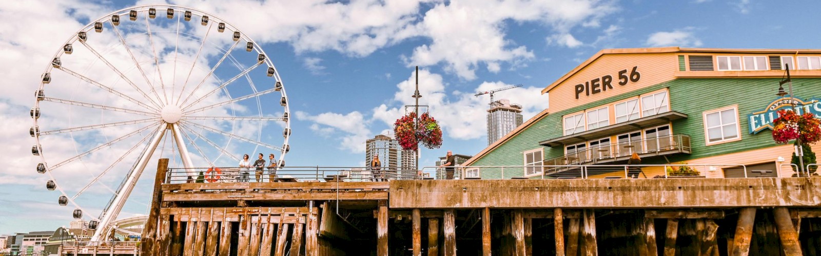 A waterfront pier with weathered pilings, a Ferris wheel in the background, a green pier building labeled “PIER 56,” and a partly cloudy blue sky.