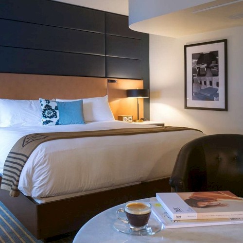 A modern hotel room with a large bed, blue headboard, nightstands, a round coffee table, stacked books, a cup, and a black chair in a cozy, well-lit space.