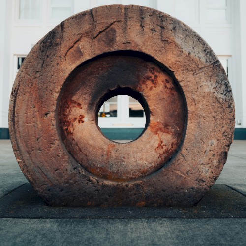 A weathered circular stone sculpture with a hollow center sits on a flat concrete surface outside a white building, framing the distant doorway.