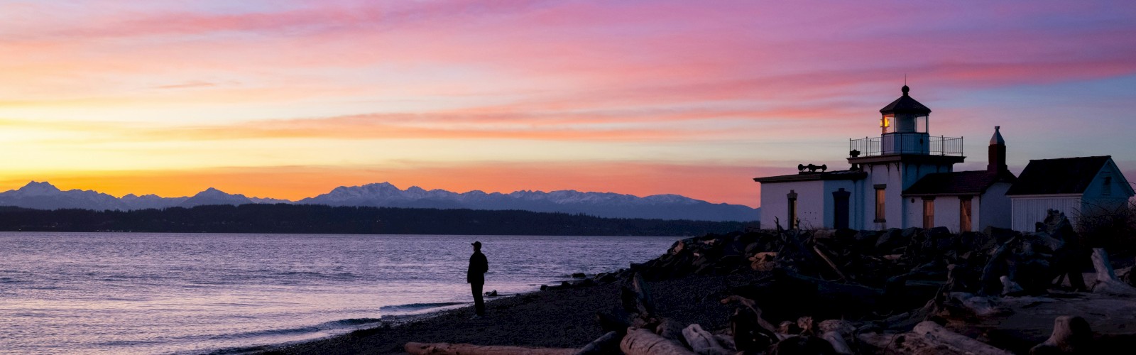 A serene coastal sunset with pink and orange skies, calm water, rocky shore, a lone figure walking, and a small lighthouse by the breakwater.