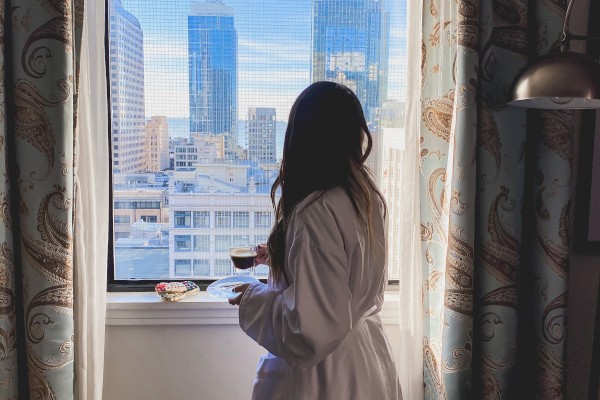 A person in a white robe stands by a hotel room window, looking out at a cityscape with tall, glassy buildings beneath a blue sky.