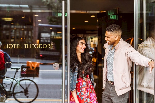 A stylish couple exits a hotel lobby and smiles at each other; a bicycle and reflections appear in the glass doors of Hotel Theodore.