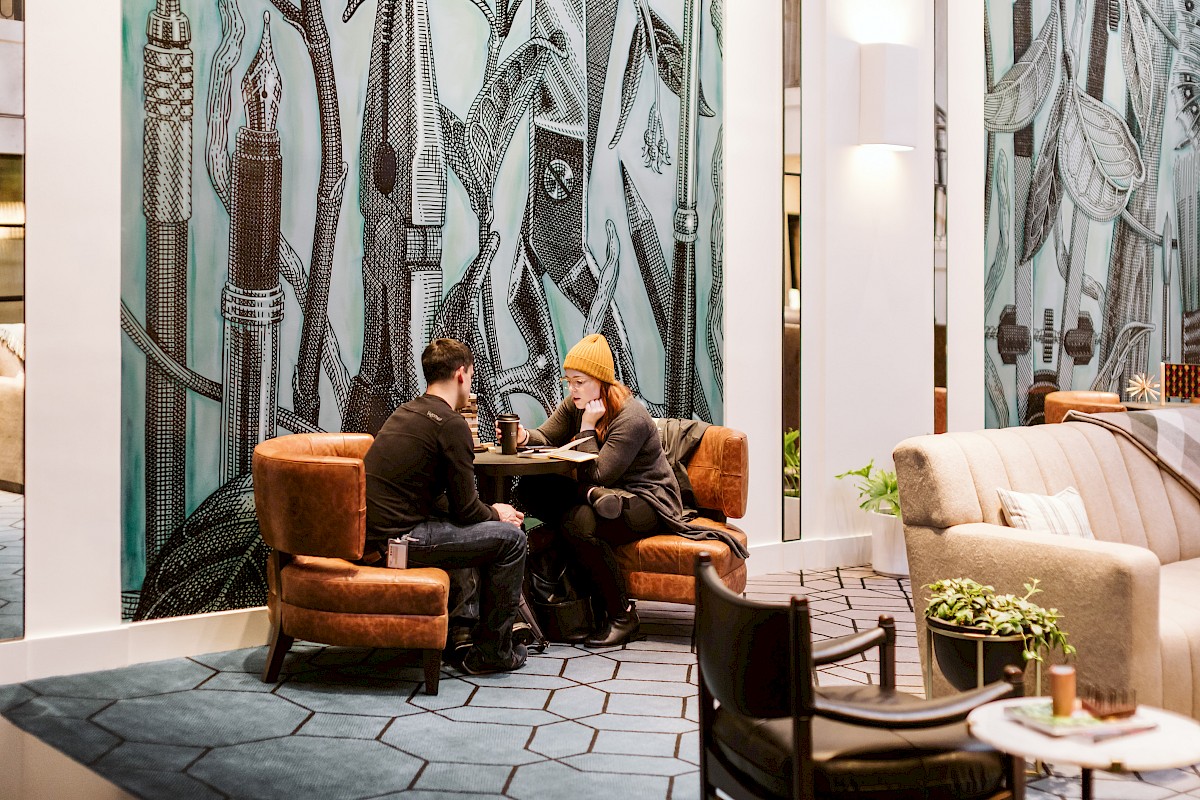 Two men sit and chat at a round table in a stylish lobby with modern seating, patterned wall art, and cozy brown and beige chairs.