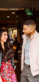 A smiling woman in a colorful dress and denim jacket greets a cheerful man in a pink jacket at the hotel entrance.