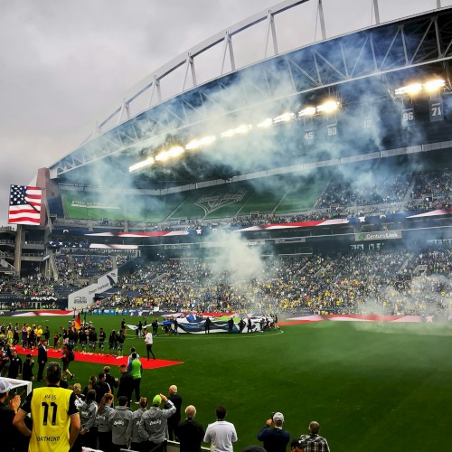 The image shows a crowded soccer stadium with smoke and flags, likely during a game or celebration, with fans in the stands.